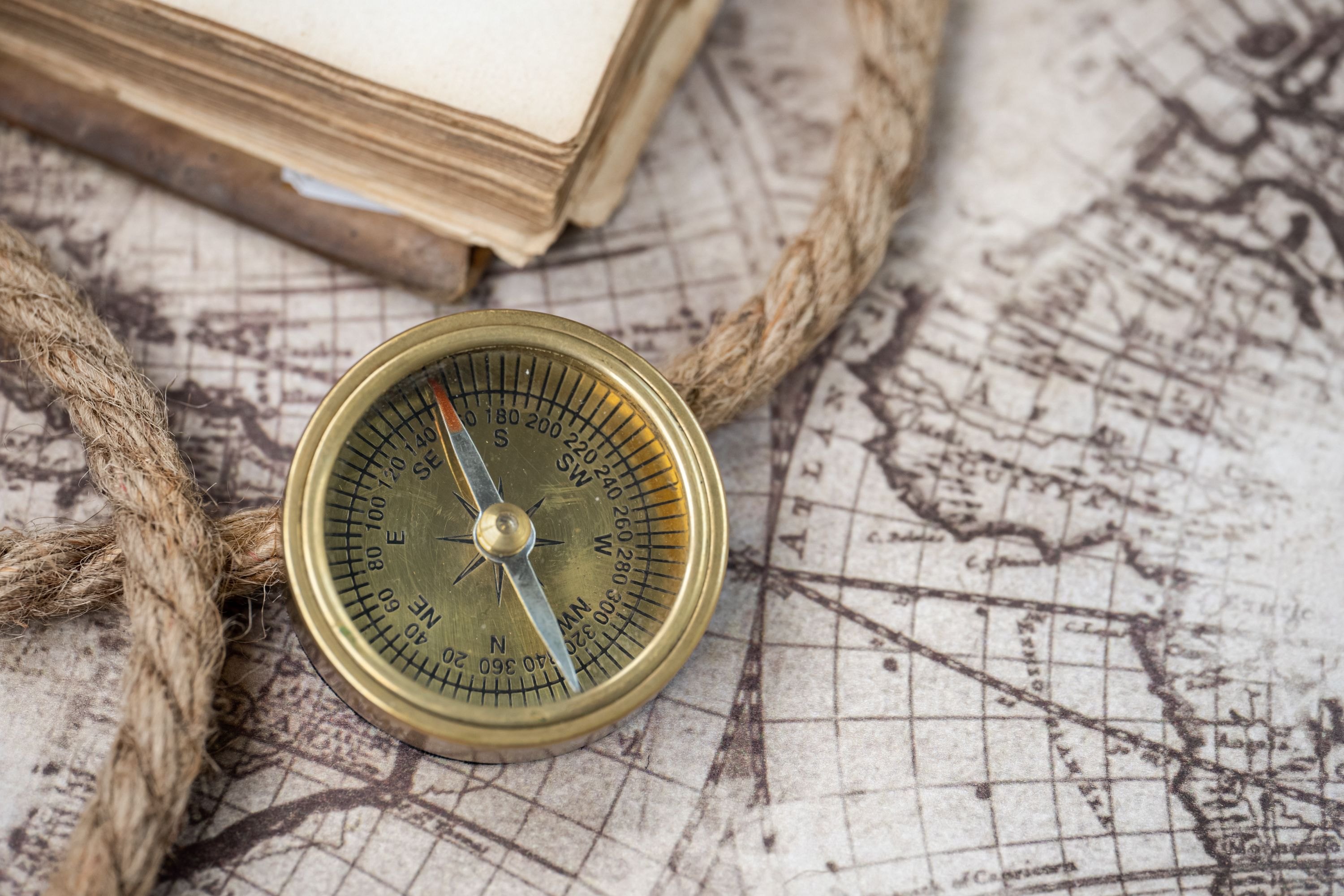 A brass compass and a coil of rope on an old map, the corner of a well-read book beside them.