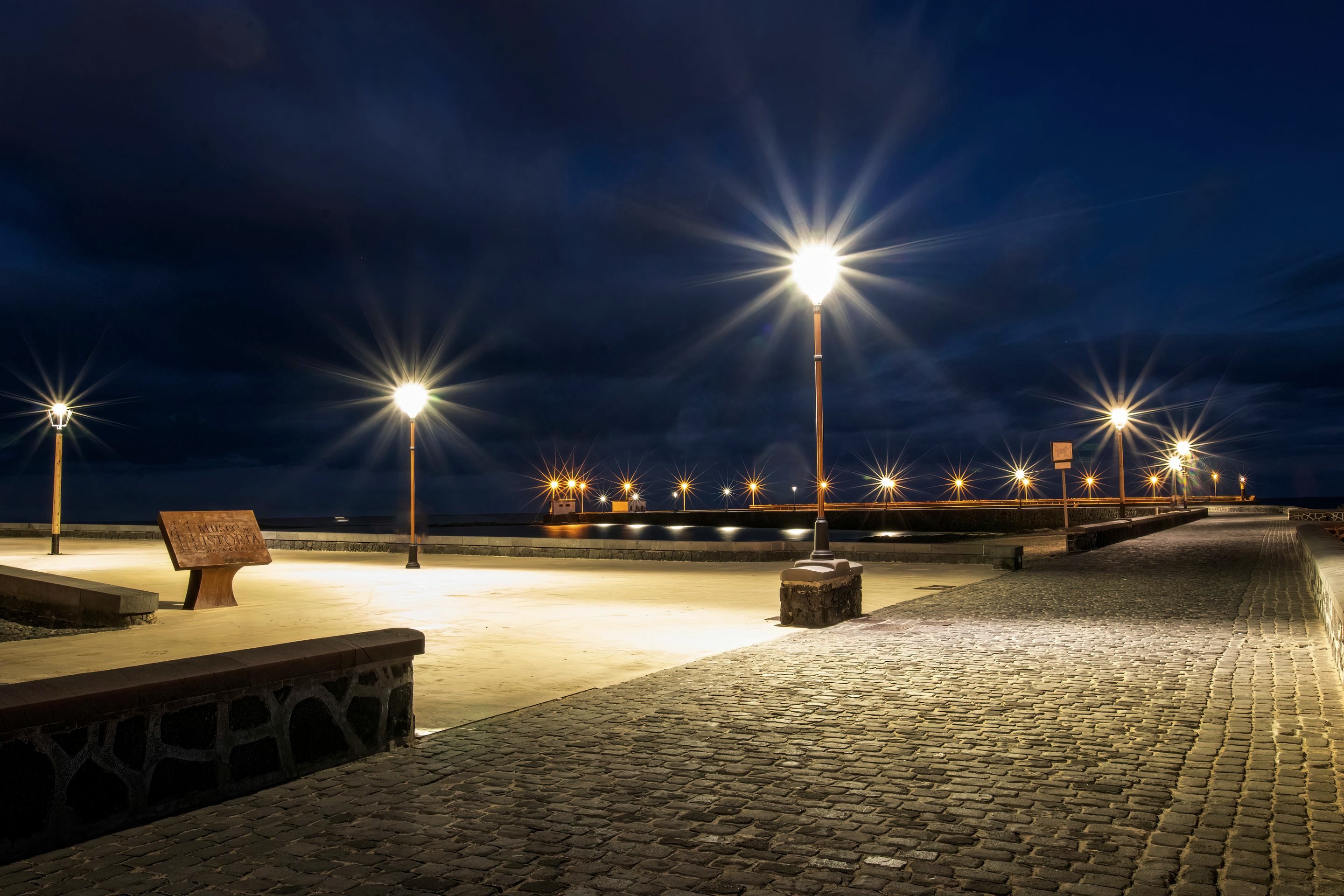 An empty cobbled seafront promenade at blue hour, lit by warm street lamps.