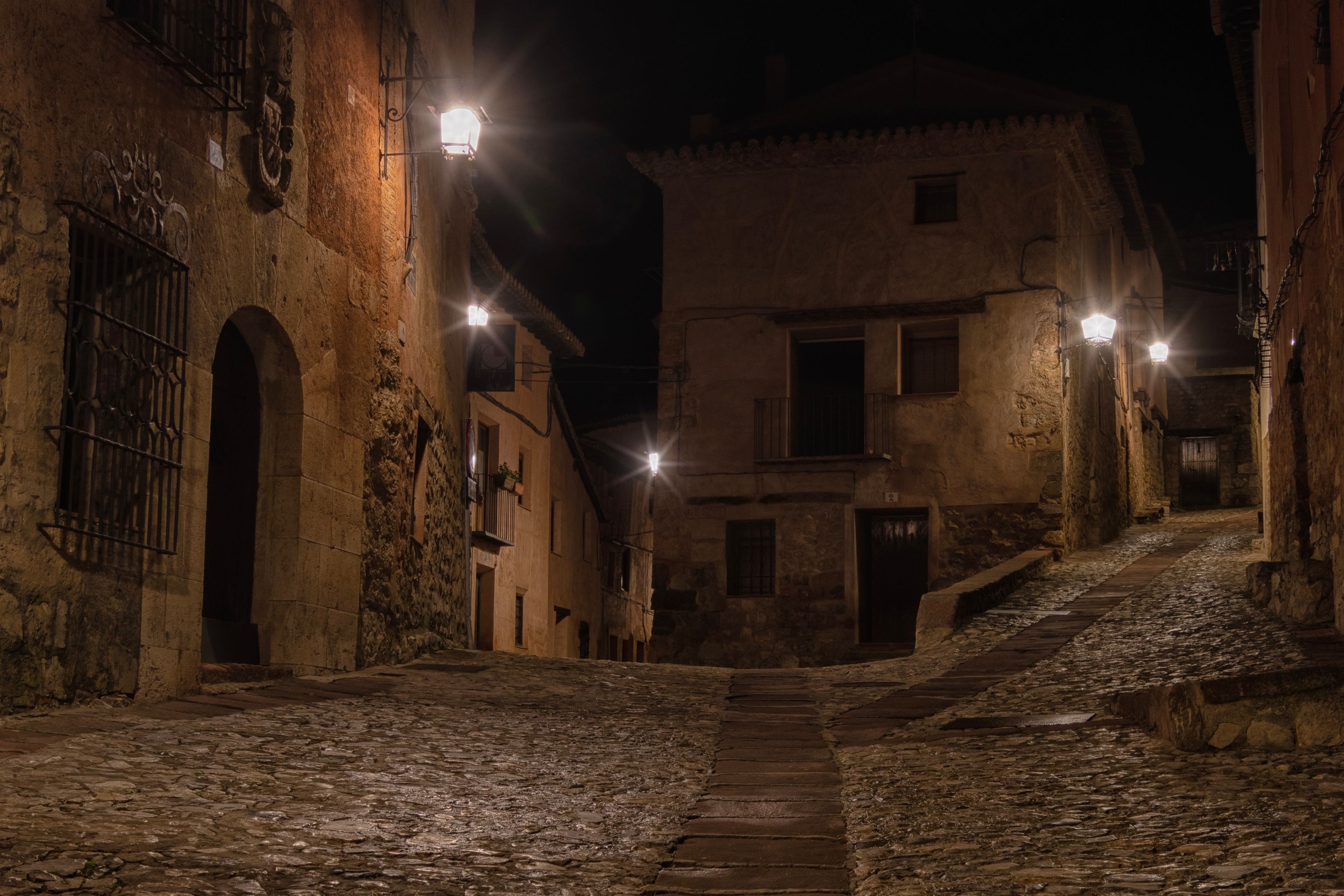 A quiet cobblestone street in a Spanish old town at night, lit by warm wall lanterns. No shop signs.