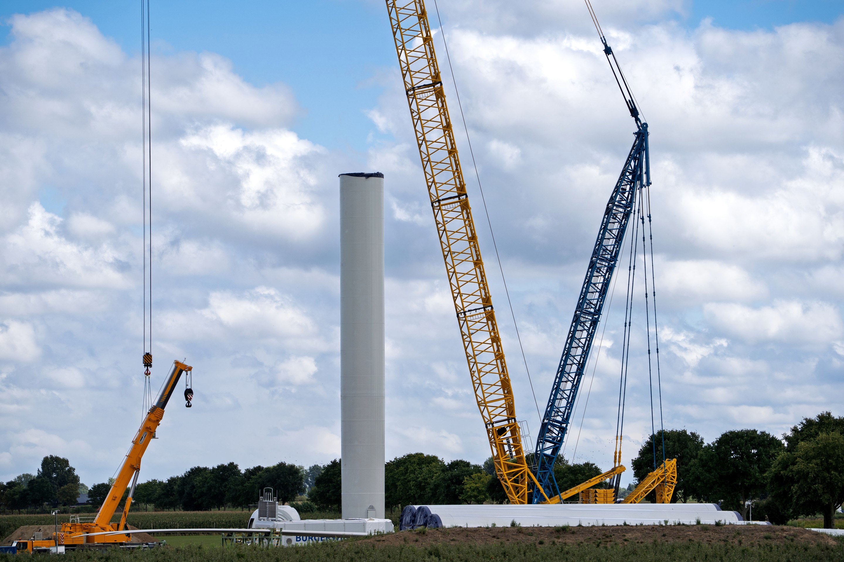 Cranes beside a wind turbine tower under construction, with blades on the ground.
